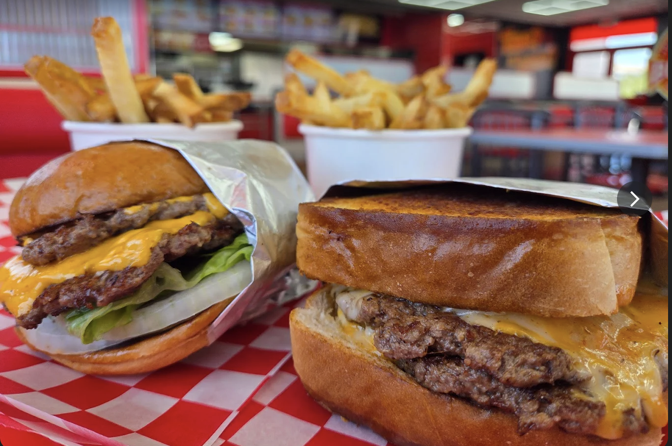 Two freshly smashed cheeseburgers with caramelized edges on red checkered paper beside cups of fries.