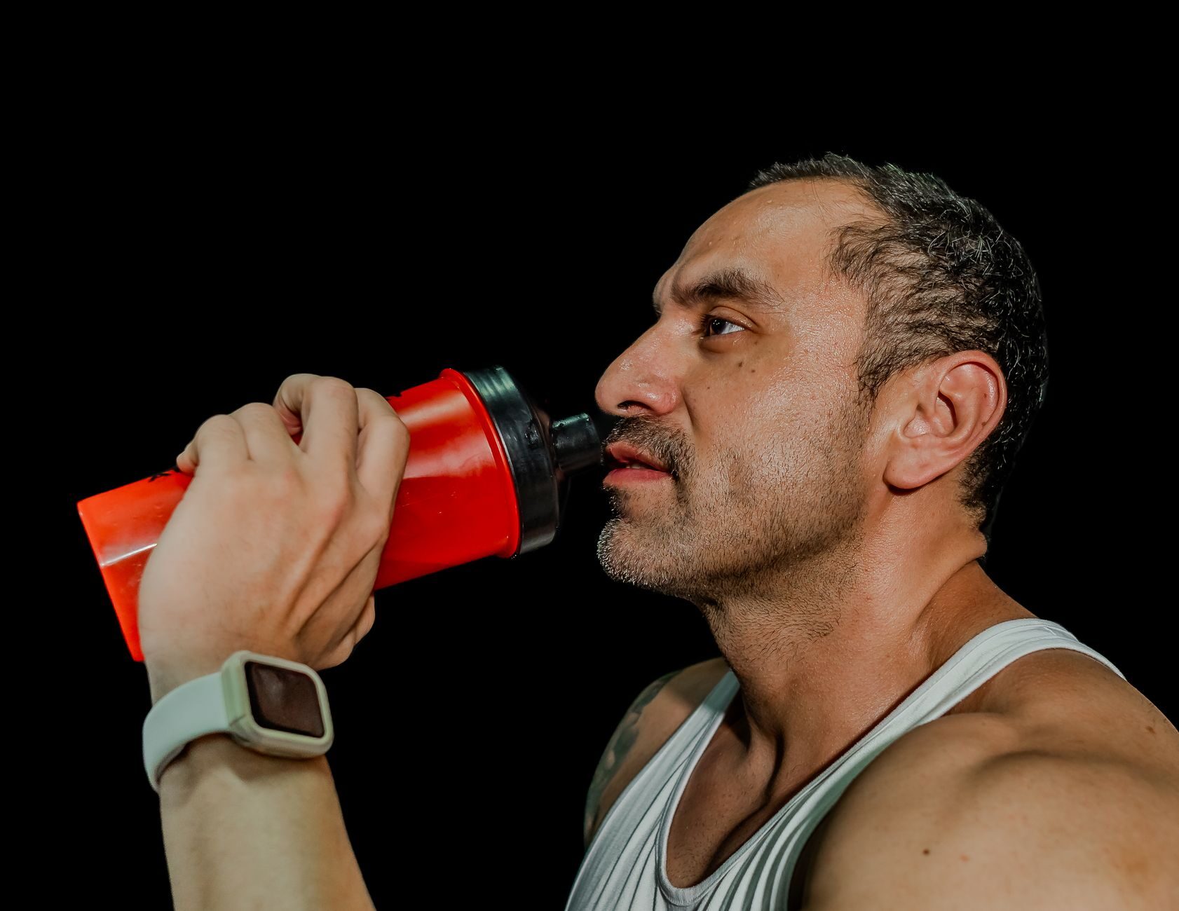 A man drinking from a red gym shaker bottle against a black background.
