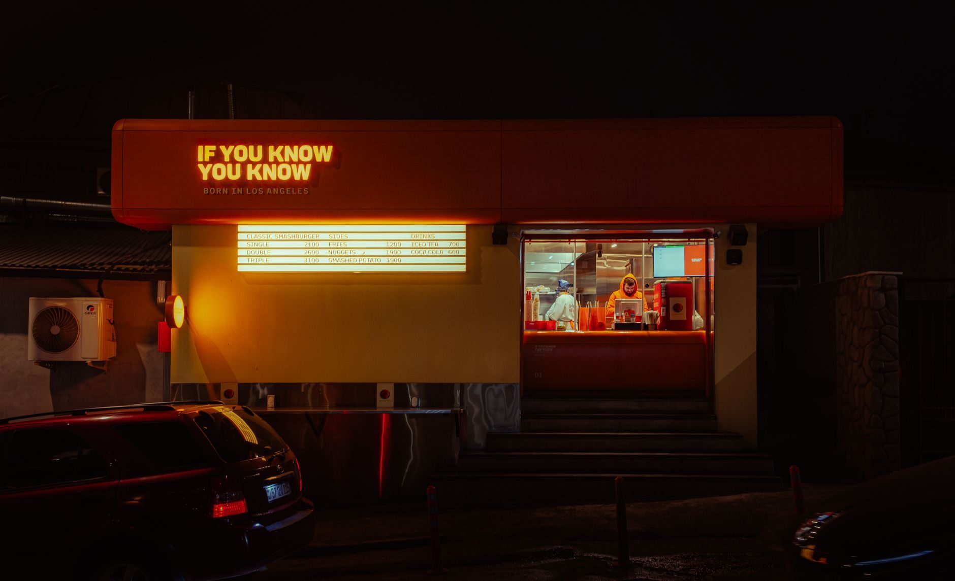 A warmly lit fast-food service window glowing against a dark parking lot at night.