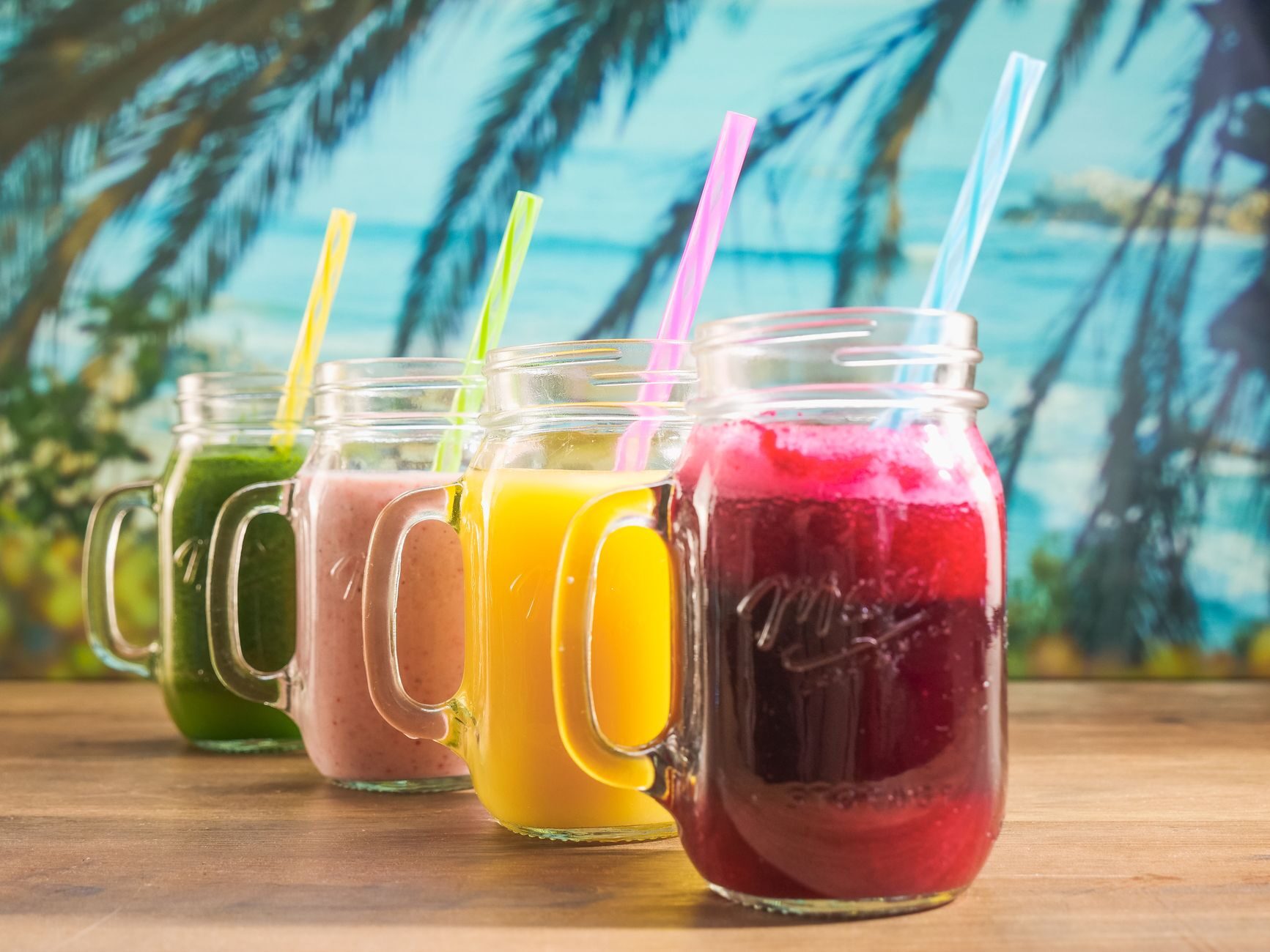 Four vivid fruit smoothies in mason jars with colorful straws.